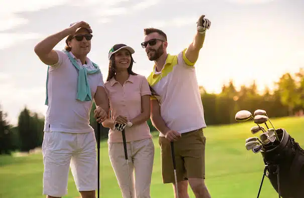 Two men and a woman are holding golf clubs, talking and smiling while standing on golf course